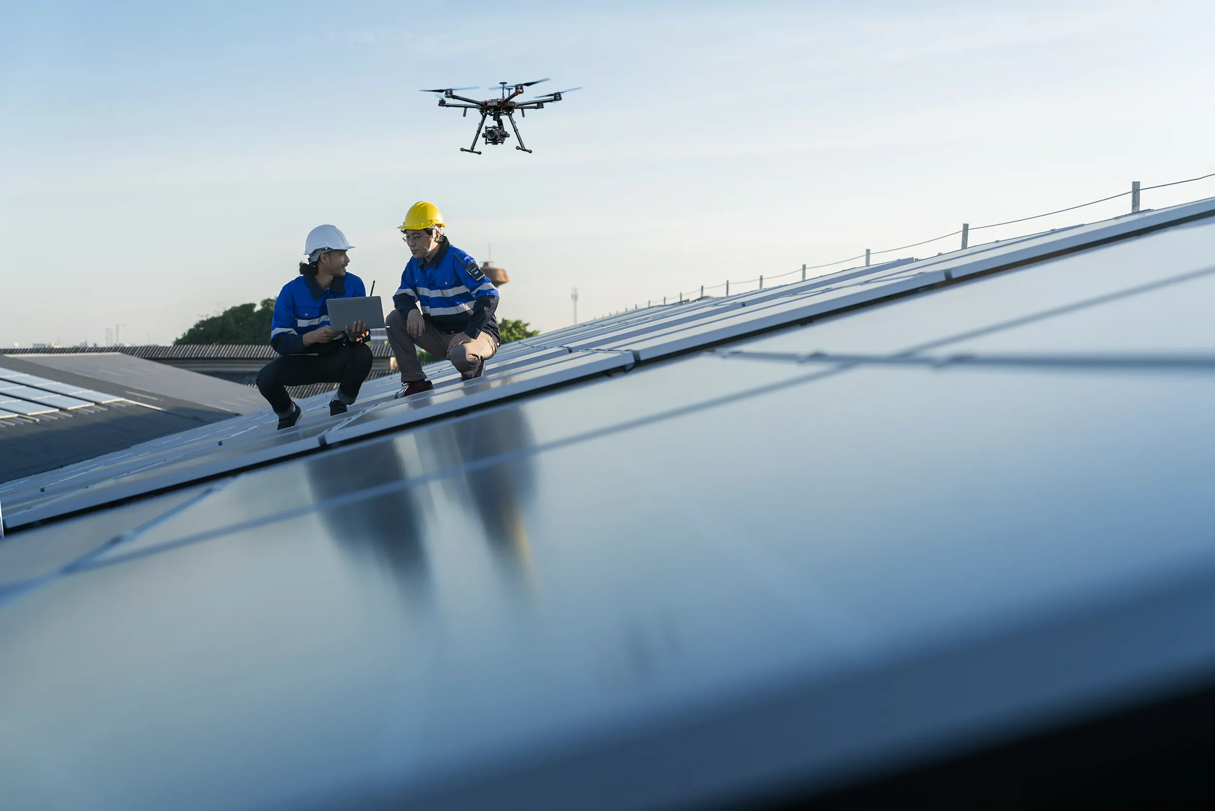 Forced perspective angle of two men kneeling on top of solar panels while a drone hovers nearby.
